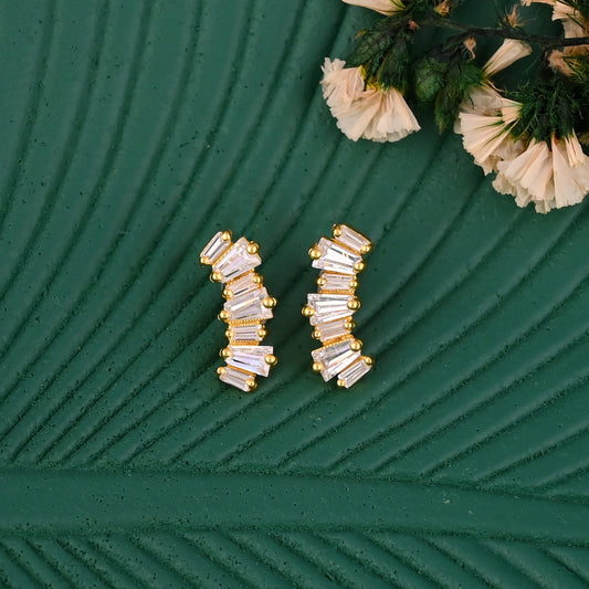 Gold and clear baguette stud earrings on a textured green surface with flowers.