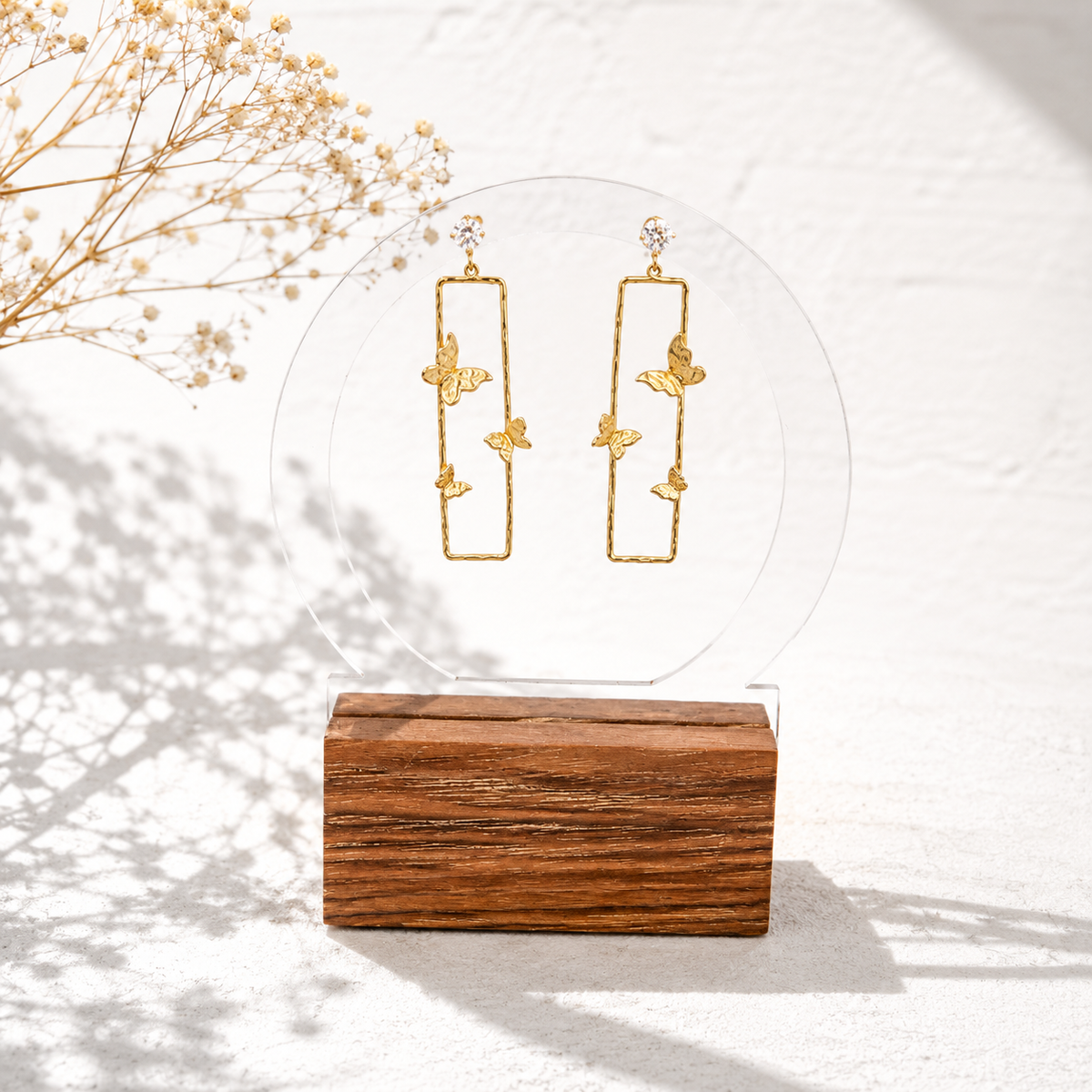 A pair of gold-toned rectangular dangle earrings with small butterfly motifs and clear stone studs, displayed on a clear circular stand over a wooden block against a white textured background.