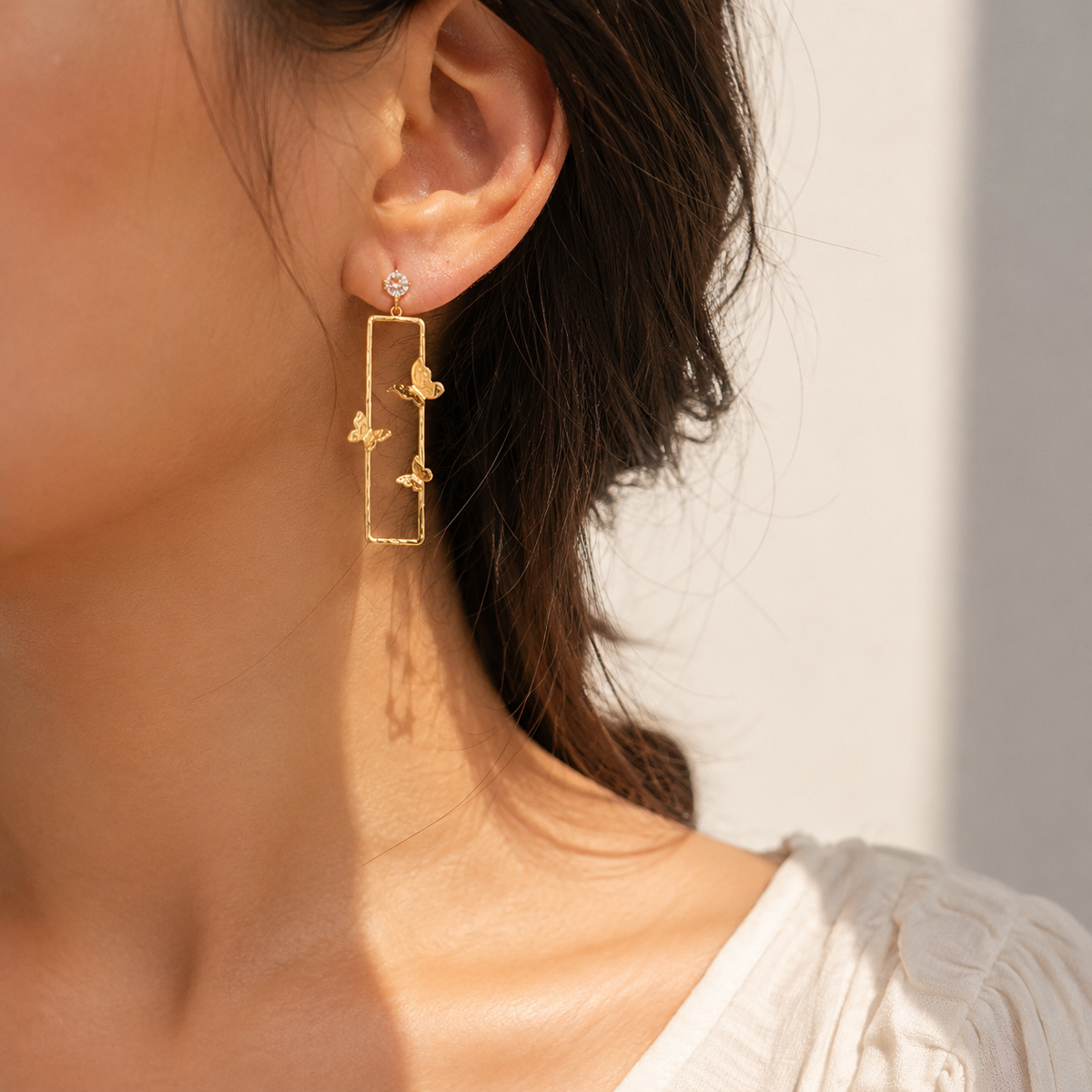 A close-up of a woman's ear wearing a delicate gold dangle earring with a rectangular frame and small floral charms, set against a soft, warm neutral background.