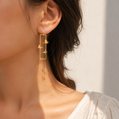 A close-up of a woman's ear wearing a delicate gold dangle earring with a rectangular frame and small floral charms, set against a soft, warm neutral background.
